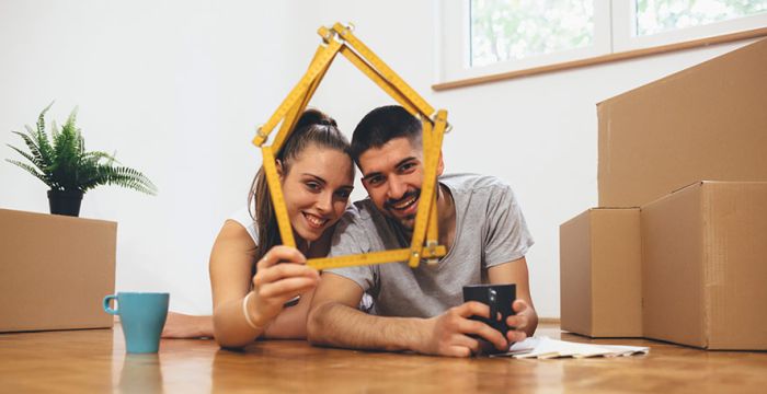 Young couple in new home with moving boxes holding measuring stick and drinking from mugs.