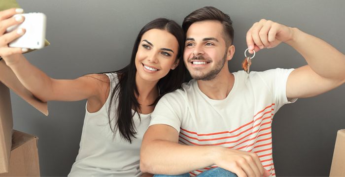 Young couple in new home with moving boxes holding keys and taking a selfie.